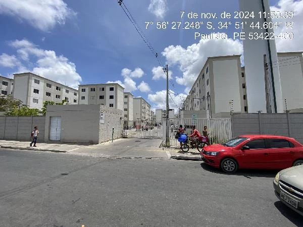 Apartamento branco em condomínio residencial com vários blocos, portaria e carros estacionados na rua em dia ensolarado