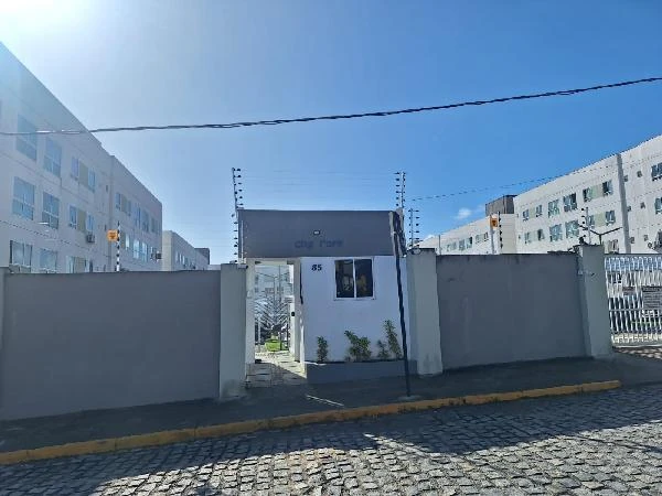 Apartamento em condomínio residencial com portaria visível e cerca elétrica em área urbana, fachada branca e muro cinza
