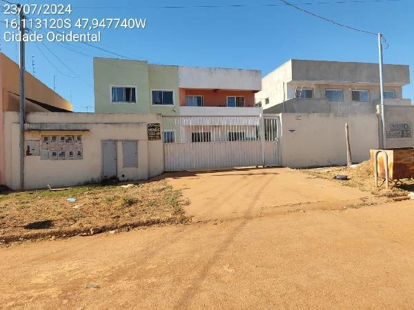 Apartamento em condomínio com fachada branca e amarela, portão branco e terreno sem pavimentação em frente