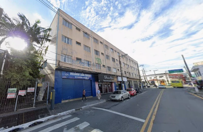 Apartamento bege antigo com janelas retangulares, localizado em rua urbana com comércio e carros estacionados