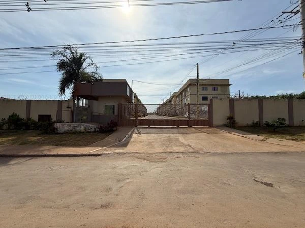 Apartamento em condomínio residencial com portão metálico, guarita e fachada clara, em área urbana com rua de terra