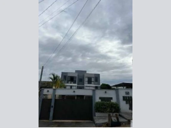 Apartamento cinza em condomínio residencial com portão preto e muros brancos em área urbana sob céu nublado