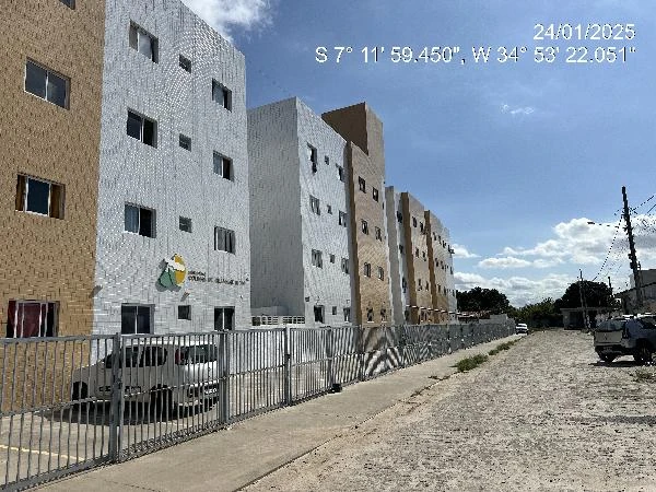 Apartamento branco e bege em condomínio residencial com cerca frontal e veículos estacionados em rua de terra