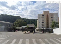 Apartamento branco em condomínio com cinco andares, vaga descoberta e área de entrada pavimentada sob céu nublado