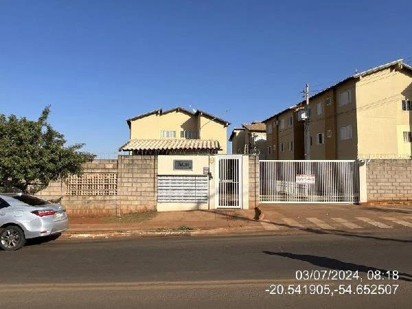 Apartamento em condomínio residencial com fachada amarela e portão branco, muro de blocos de concreto e calçada asfaltada