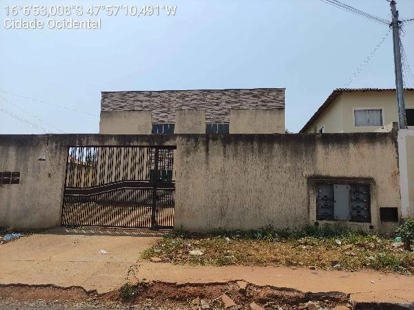 Apartamento branco com muro alto e portão de ferro em área residencial com calçada danificada