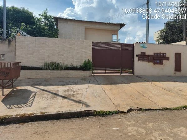 Apartamento com fachada clara, portão de metal marrom e muro baixo em área urbana sob céu parcialmente nublado