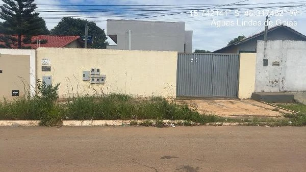 Apartamento com parede amarela e portão cinza em rua residencial com vegetação alta e casas ao fundo