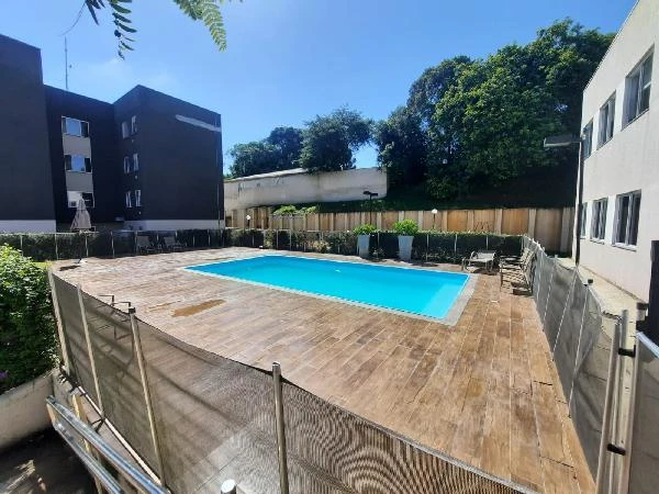 Apartamento de piscina azul cercada por tela, prédio de fachada simples com janelas e área externa com árvores ao fundo