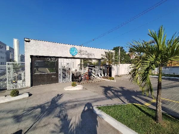 Apartamento em condomínio residencial com portaria, área externa pavimentada e vegetação ao redor, fachada clara e moderna