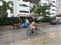 Apartamento branco de 4 andares com área externa comum, veículos estacionados e ciclista em via molhada