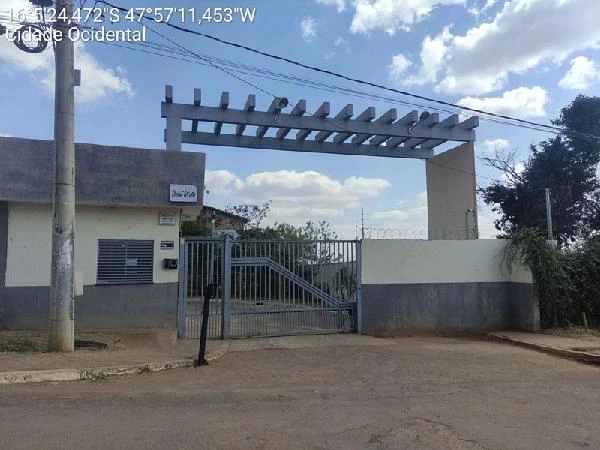 Apartamento em condomínio residencial com portão metálico e estrutura cinza em área urbana sob céu parcialmente nublado