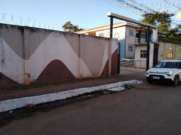 Apartamento em condomínio residencial com fachada branca e bordo, muro com grades de proteção e carro branco estacionado na rua