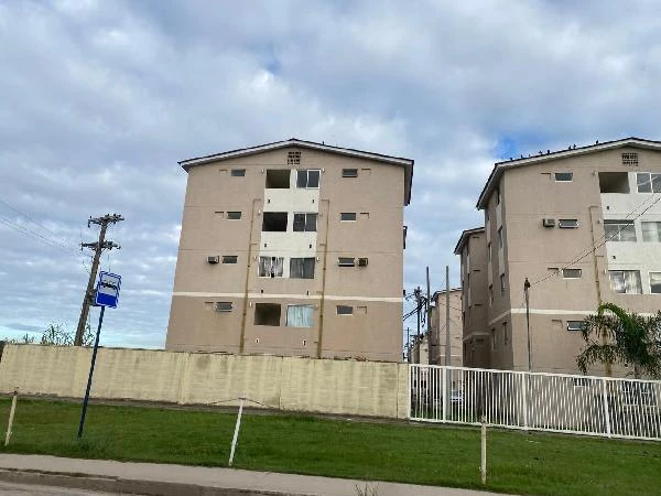 Apartamento bege com 4 andares em condomínio com muro baixo e portão branco visto de frente em área urbana com céu nublado