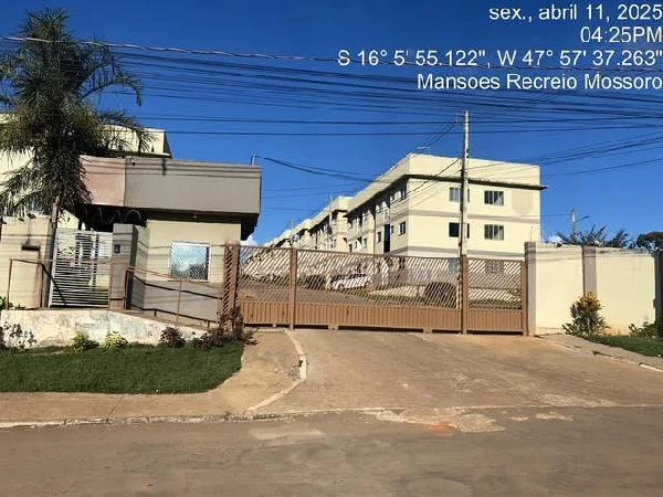 Apartamento com fachada de condomínio residencial branco e portão automático em rua asfaltada com palmeira próxima