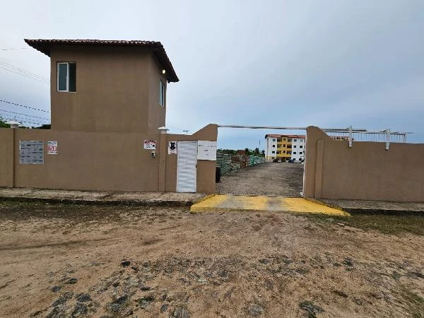 Apartamento bege em condomínio com torre de vigilância na entrada e área com edifício ao fundo em terreno aberto