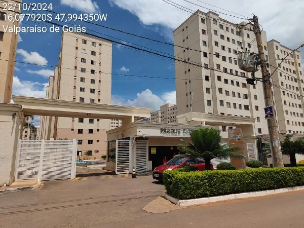 Apartamento em condomínio residencial com fachada clara, portaria de entrada, veículos estacionados e área comum externa visível