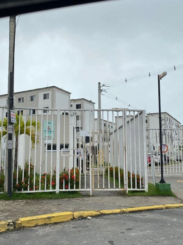 Apartamento branco em condomínio fechado com portão metálico branco e prédios residenciais ao fundo.