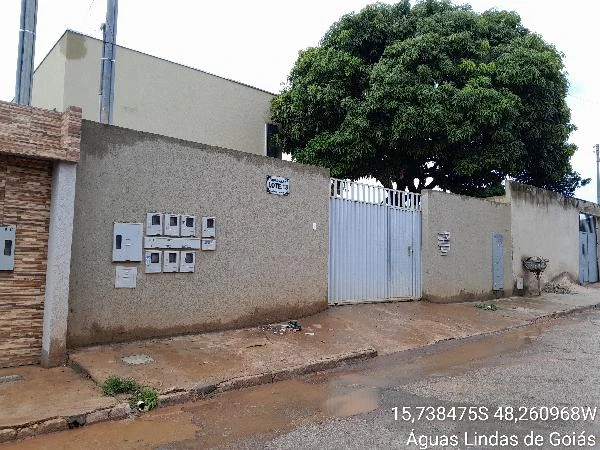 Apartamento no condomínio com muro cinza, portão metálico branco e árvore ao fundo na rua pavimentada