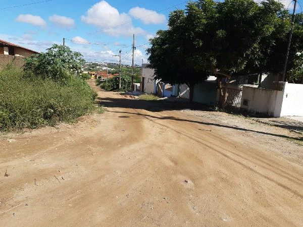 Terreno e rua de terra com casas residenciais ao lado em área urbana com vegetação e céu claro