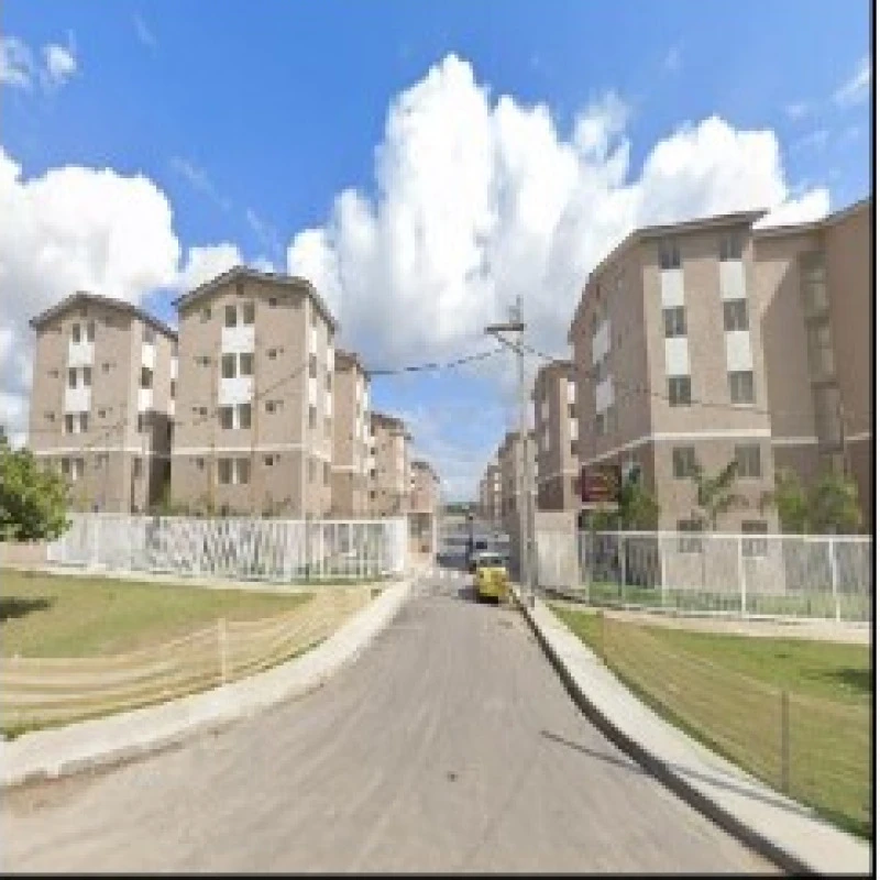 Apartamento bege em condomínio residencial com múltiplos blocos de 4 andares e ruas asfaltadas com cerca branca e céu azul nublado.