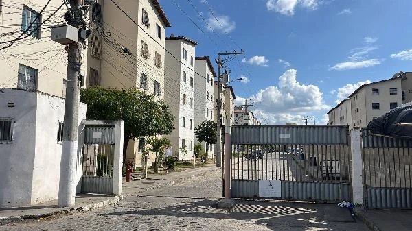 Apartamento em condomínio residencial com portaria, prédios de fachada clara e área comum com árvores, sob céu azul e nuvens
